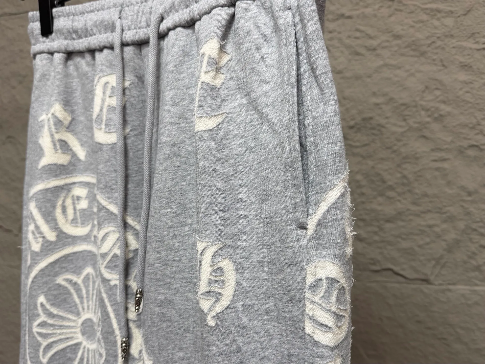 CH/Cro Heart 2026ss Burnt Flower Straight-leg Sweatpants Classic heavy-weight sweatpants with gray tones, Retro street style full-fitting, direct recognition, same style for men and women. ●Three-dimensional patchwork technology: The whole body is covered with Gothic letters + cross flower totems, using raw edge patchwork embroidery, with a mustache-like old texture, and uneven and layered patterns. It is not an ordinary print, and it is durable and not easy to fall off. ●Style tailoring: Loose, straight and wide-legged version, modifying the shape of the legs, hiding the flesh neatly, not flattering the figure, with an online drape, and the upper body has a relaxed street tone. ●Fabric and waist: Heavy knitted terry fabric, thick and soft, with good drape; elastic drawstring waist, with branded metal engraved rope ends, easy to put on and take off, and strong waist adaptability. ●Detailed design: The side pockets are convenient for carrying items, and the back pocket is decorated with cross-pattern patches. The front and rear patterns echo each other. Every detail is highly recognizable to the brand. ●Wearing scenarios: Suitable for street wear, daily outings, and trendy wear. Paired with sweaters, denim jackets, and leather jackets, you can easily create an American retro cool style. ●The overall gray base is paired with Cream White patches, which are low-key yet impactful. They are both comfortable and trendy. Versatile sweatpants, a must-have for fashionistas, are also available at the counter. SIZE：M L XL XXL XXXL